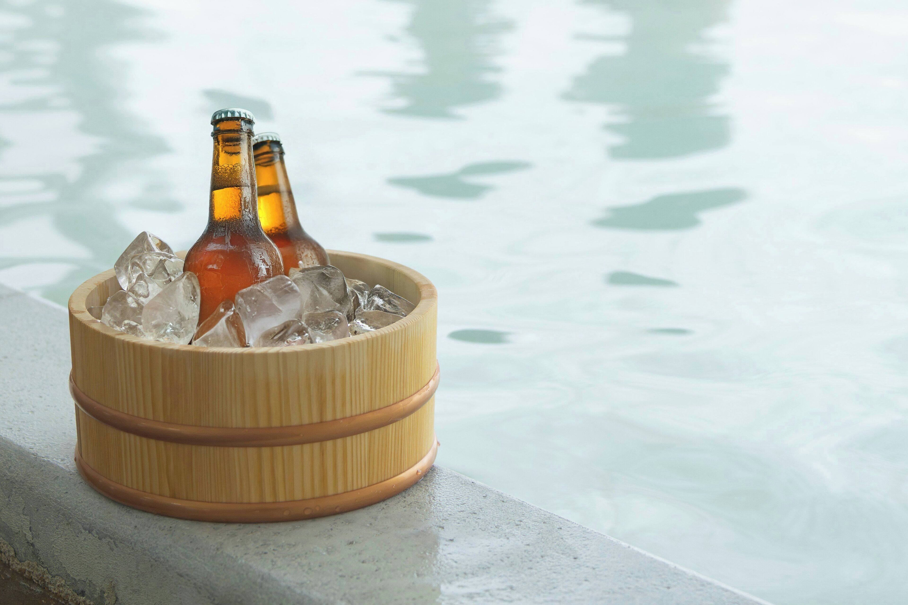 A bamboo cooler with bottles and ice cubes on a ledge by a pool.