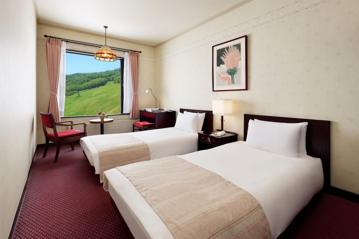 Quarto de hotel com duas camas, uma escrivaninha, uma cadeira, uma janela com vista para uma paisagem verde e um quadro na parede.