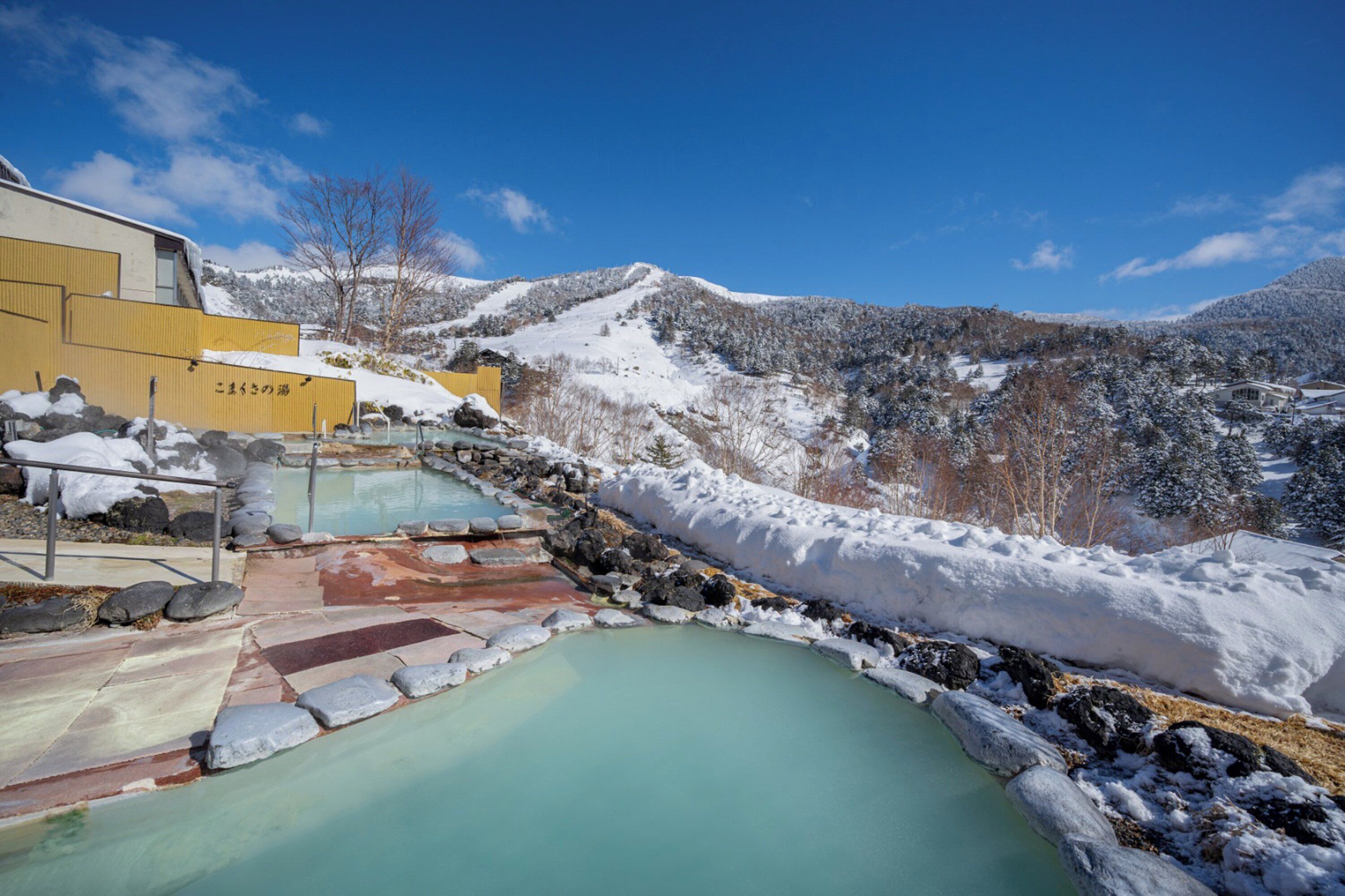 Uma área de águas termais com um caminho de pedra que leva a uma piscina, cercada por montanhas cobertas de neve.