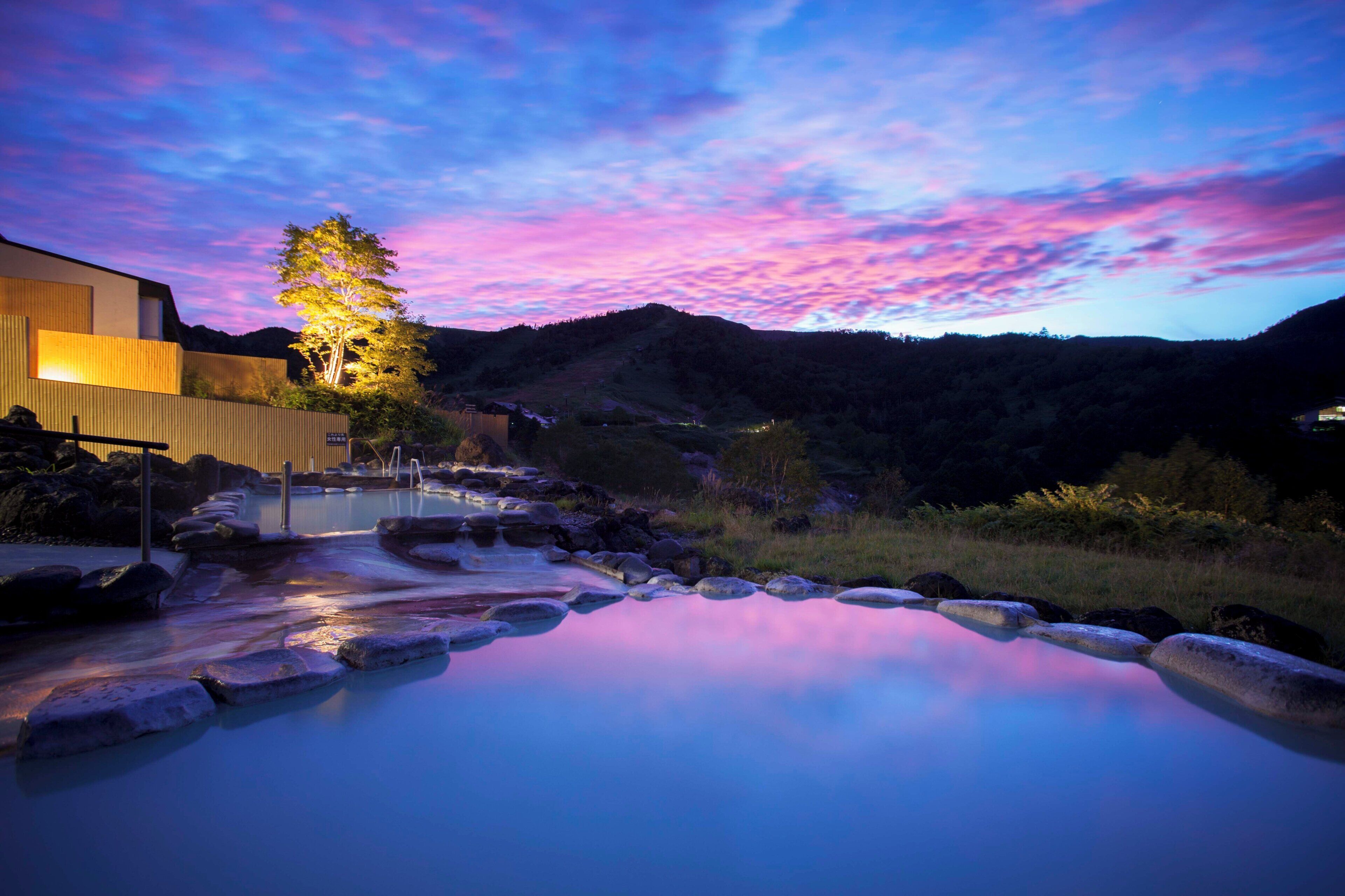 A modern building with a pool area, surrounded by natural landscape and a colorful sky.
