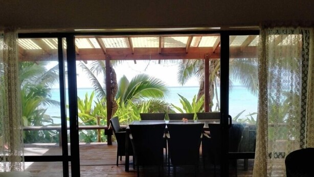 Un pontile in legno con tavolo e sedie, affacciato su una spiaggia tropicale con palme e l'oceano.