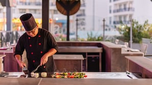 A chef preparing food at an outdoor restaurant with a view of buildings in the background. - Muthu Oura Praia Hotel (Albufeira)
