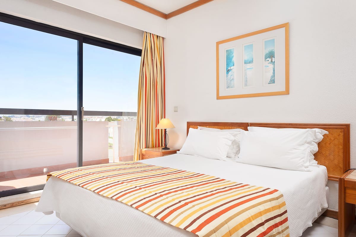 A hotel room with a large bed, a nightstand, a lamp, and a window with a view of the city.