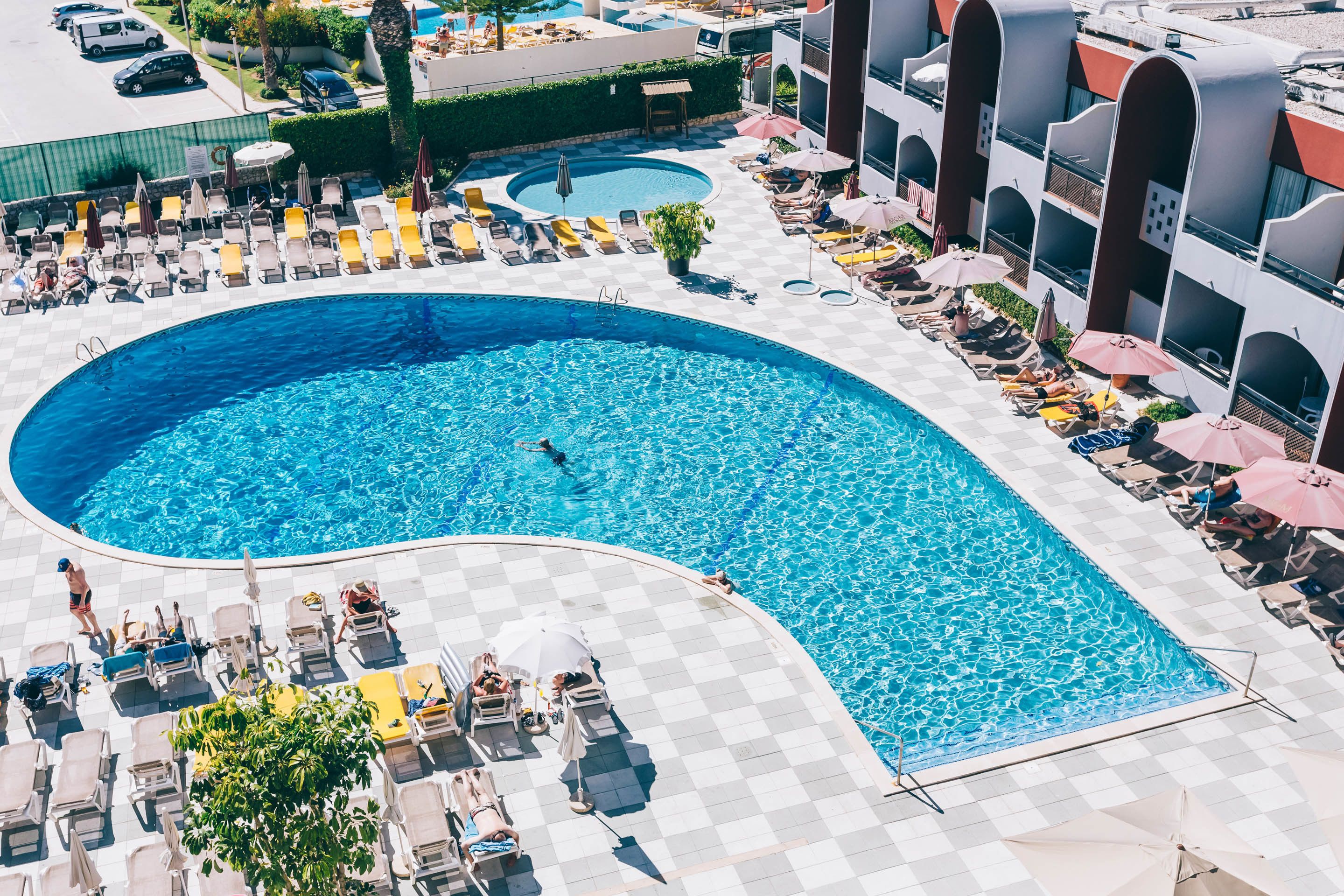 Una amplia piscina con tumbonas y sombrillas, rodeada por un solárium a cuadros y un edificio con balcones arqueados.