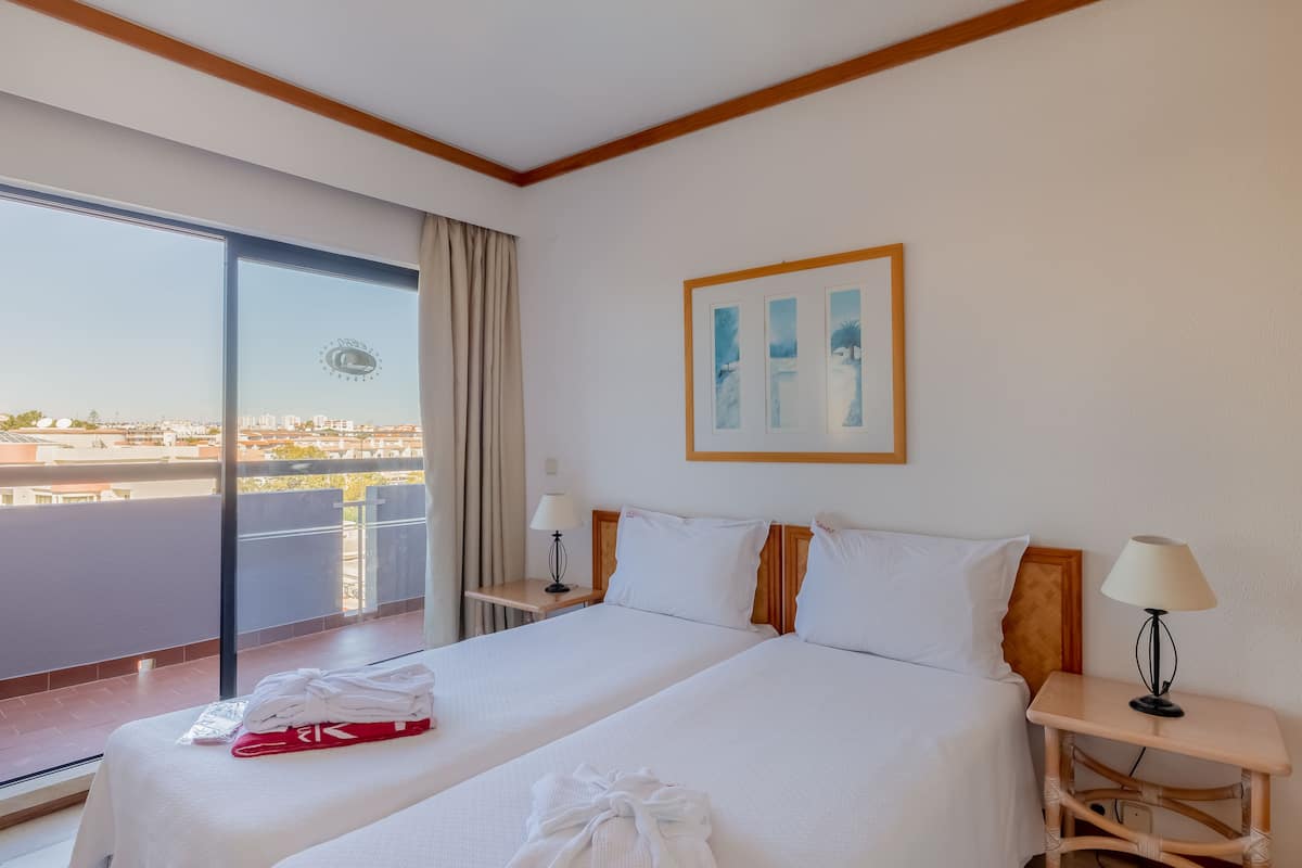A hotel room with two beds, a large window with a view, a painting on the wall, and a wooden ceiling.
