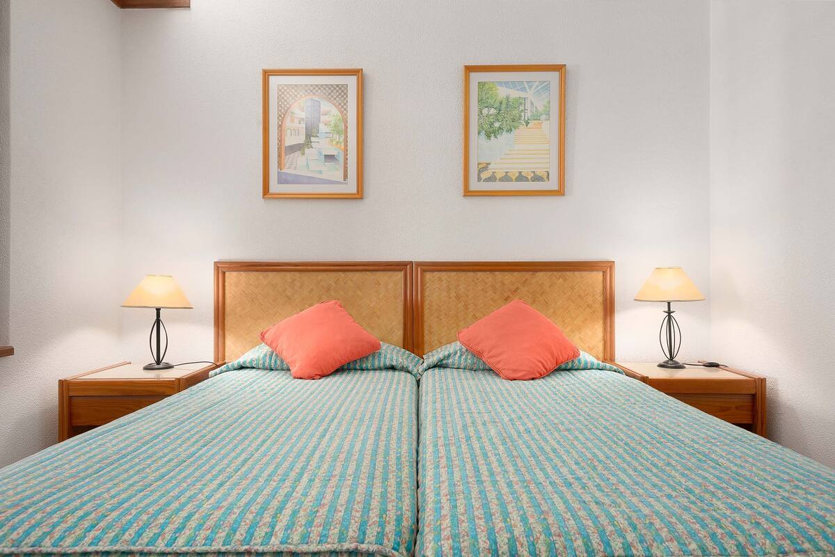 A double bed with a patterned bedspread, two nightstands with lamps, and two framed pictures on the wall.
