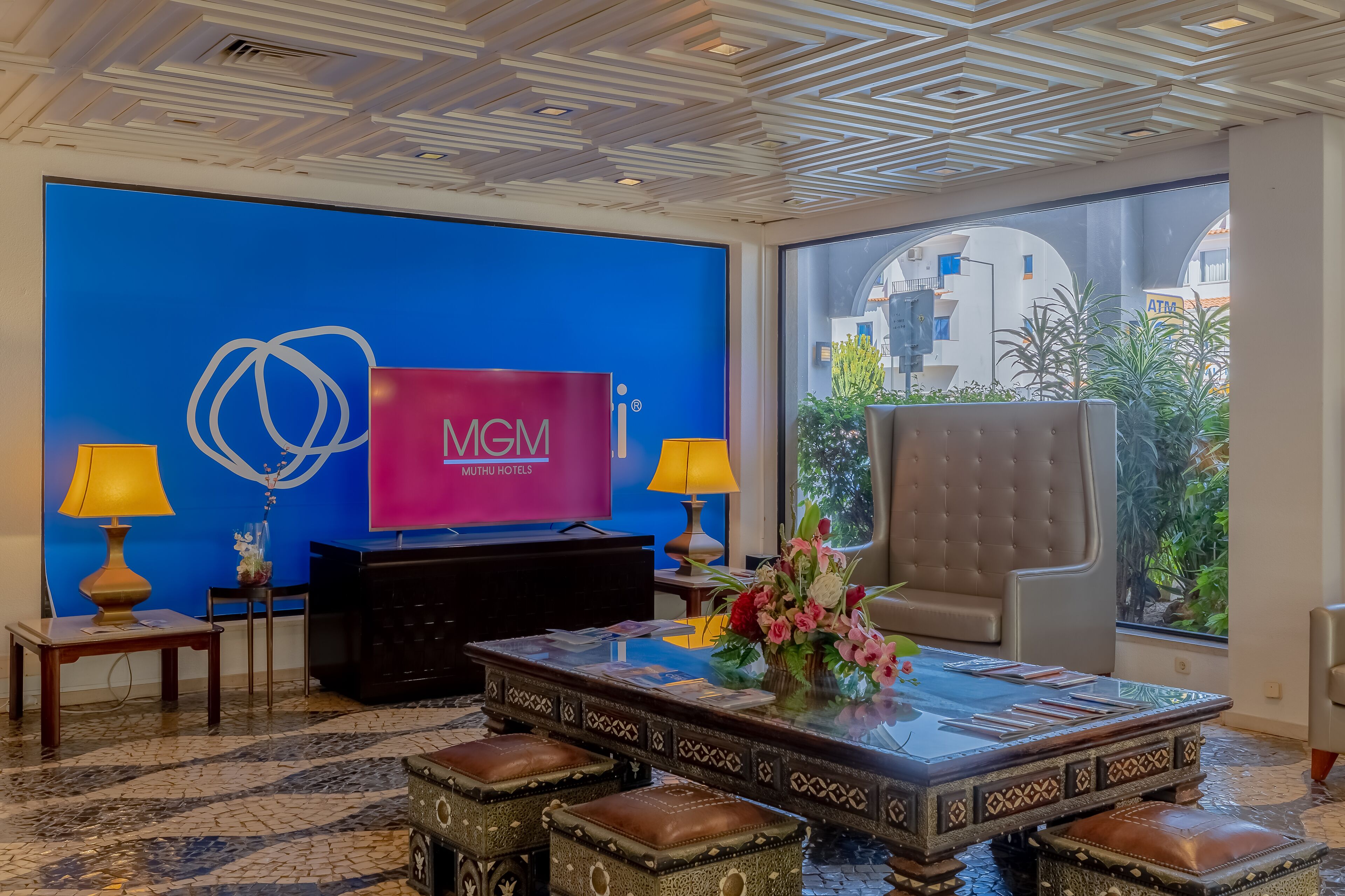 En entréhall med en blå vägg, en stor tv-skärm som visar MGM MUTHU HOTELS, en receptionsdisk, två lampor, en dekorativ vas, en fåtölj i läder och ett mönstrat soffbord.