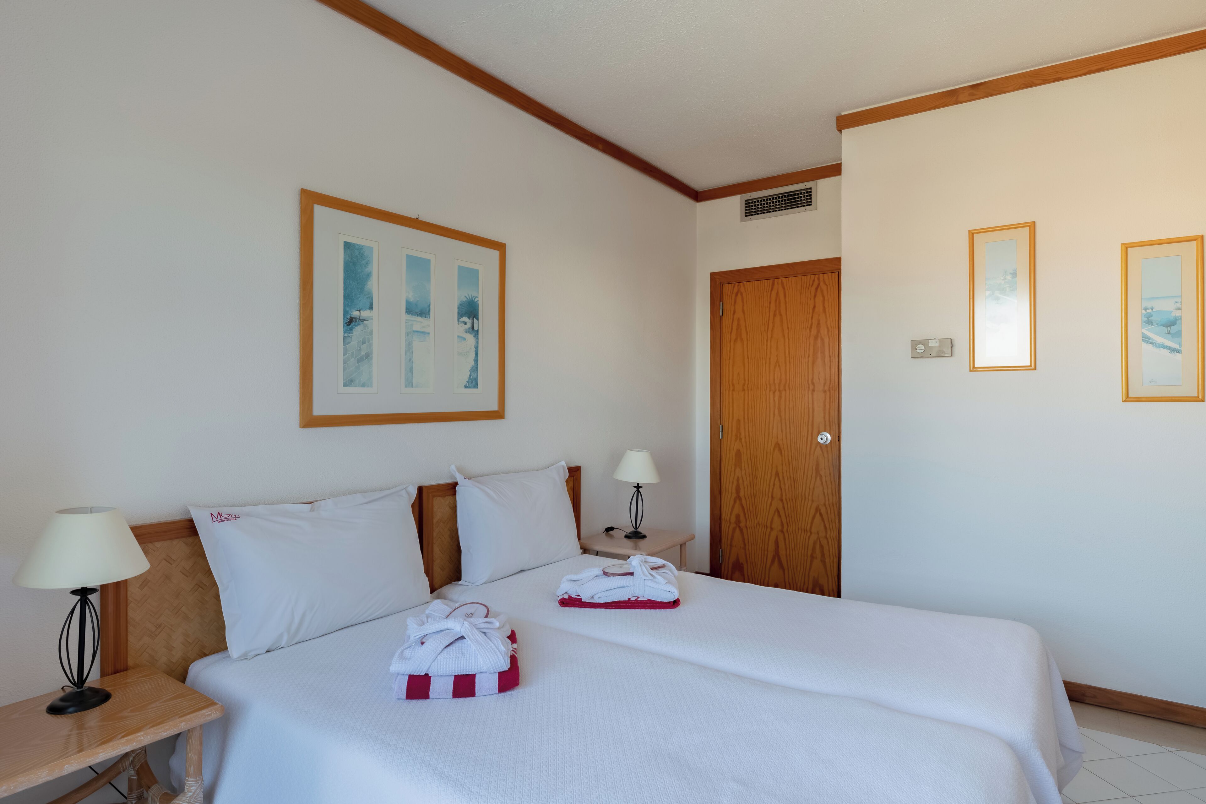 A hotel room with two beds, a wooden headboard, a nightstand with a lamp, and framed pictures on the wall.