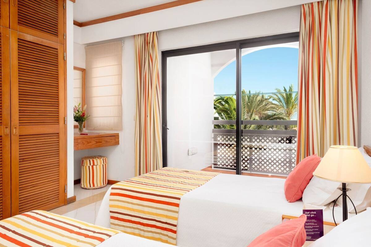 A hotel room with a bed, a desk, a chair, a lamp, and a view of palm trees.