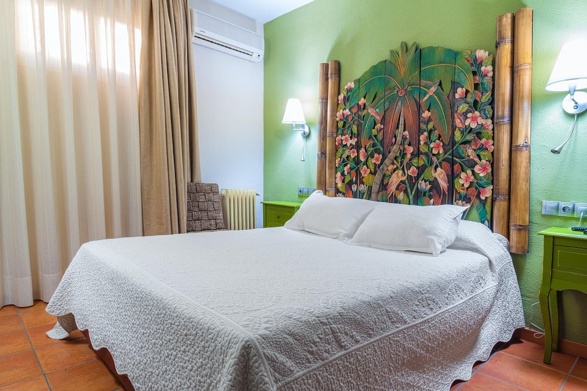 Ein ordentlich bezogenes Bett mit weißer Decke, eine grüne Wand mit floralem Wandbild, eine Lampe und ein grüner Nachttisch.