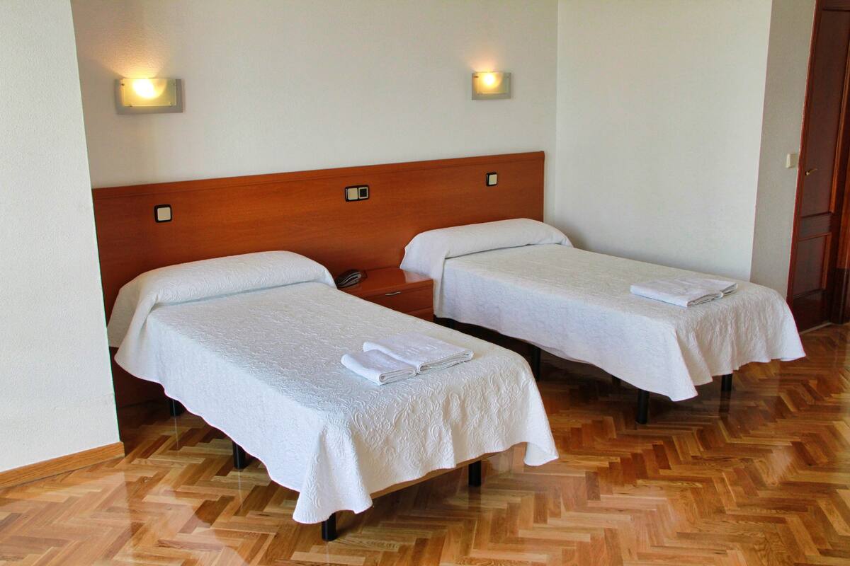 Two single beds with wooden headboards and white linens, a wooden parquet floor, and wall-mounted lights.