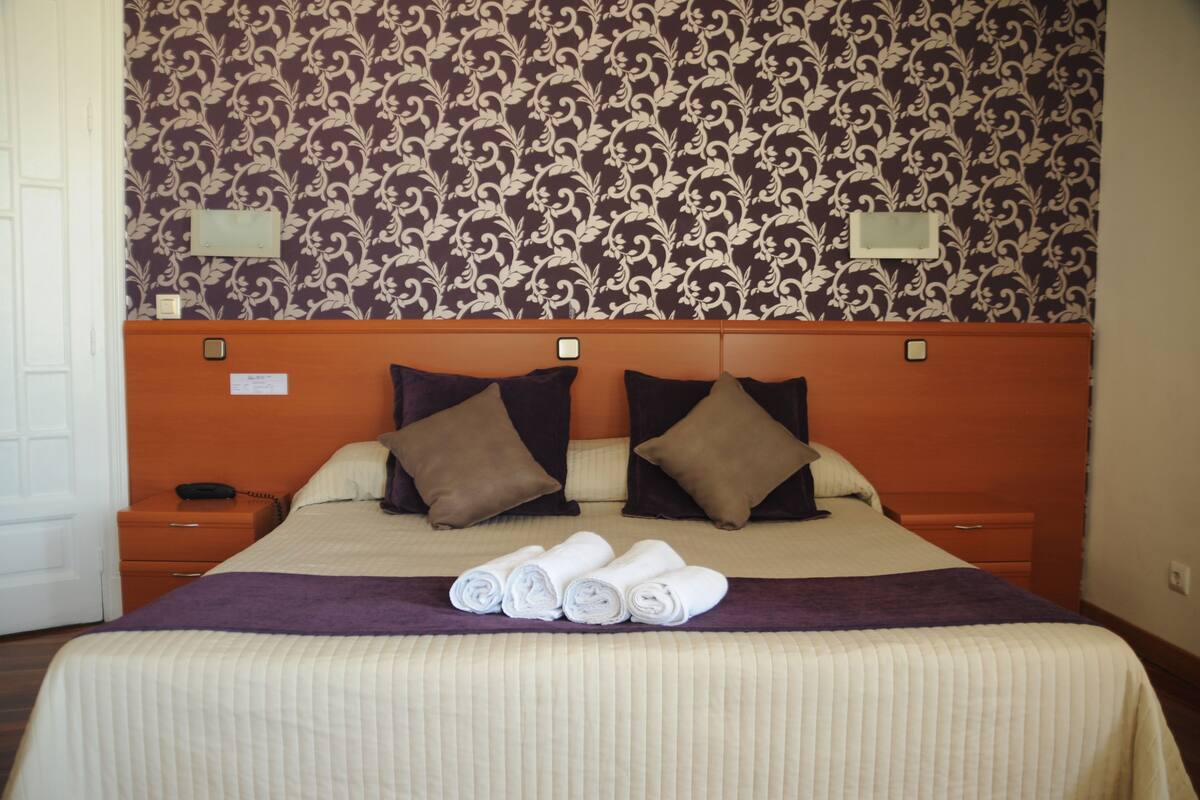A neatly made bed with rolled towels, a headboard, and a patterned wallpaper.