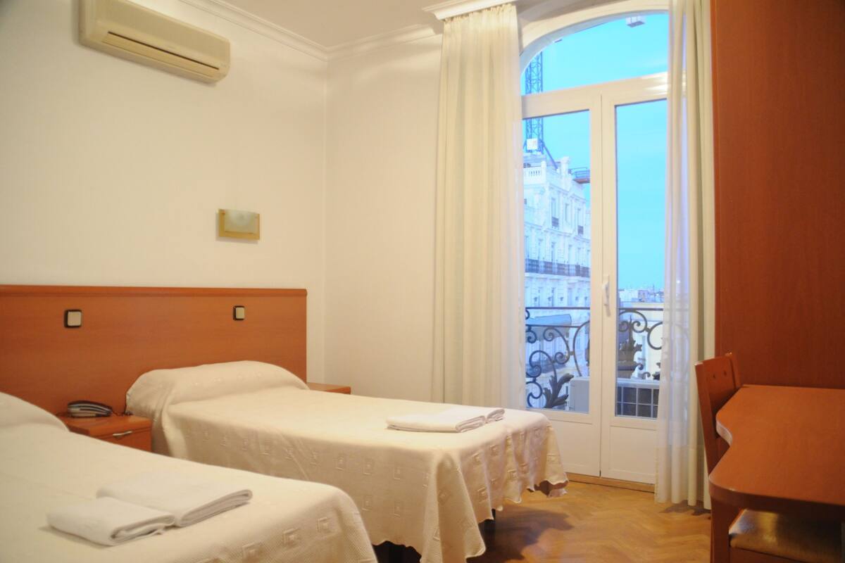 A hotel room with two beds, a small table, and a view of a balcony with a railing.