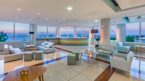 A spacious lobby with a modern design, featuring a seating area with sofas and chairs, a central coffee table, and a large window offering a view of the cityscape. - Kipriotis Aqualand by Anayia All Inclusive Resorts (Kos)