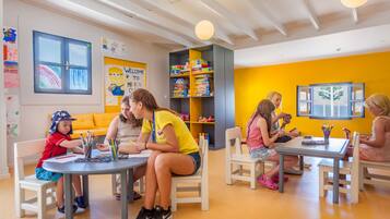 A brightly lit indoor space with children engaged in activities at tables, a bookshelf filled with books, and a yellow wall with a welcome sign.