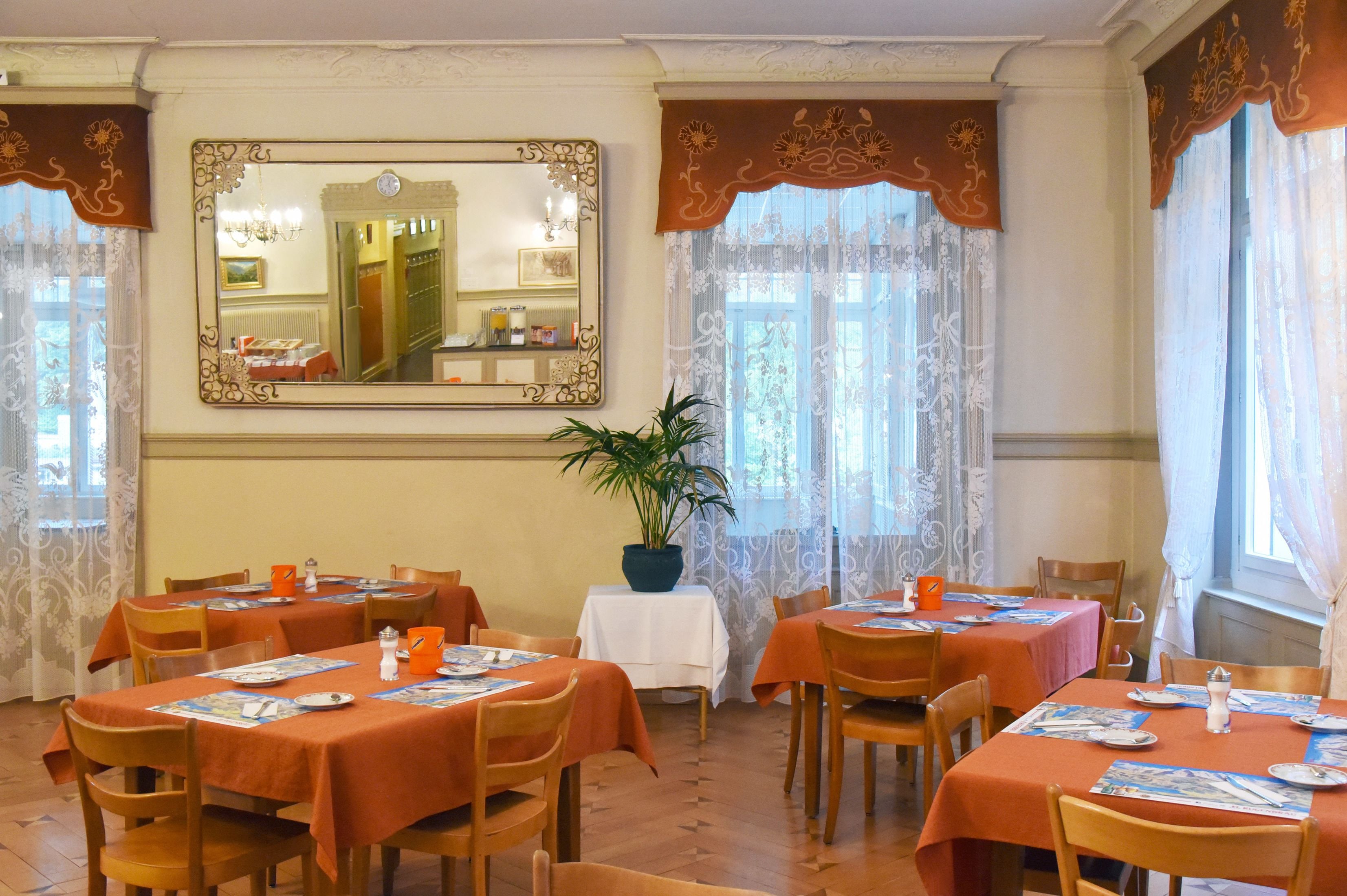 A dining area with tables set for four, a large mirror on the wall, and a potted plant in the center.