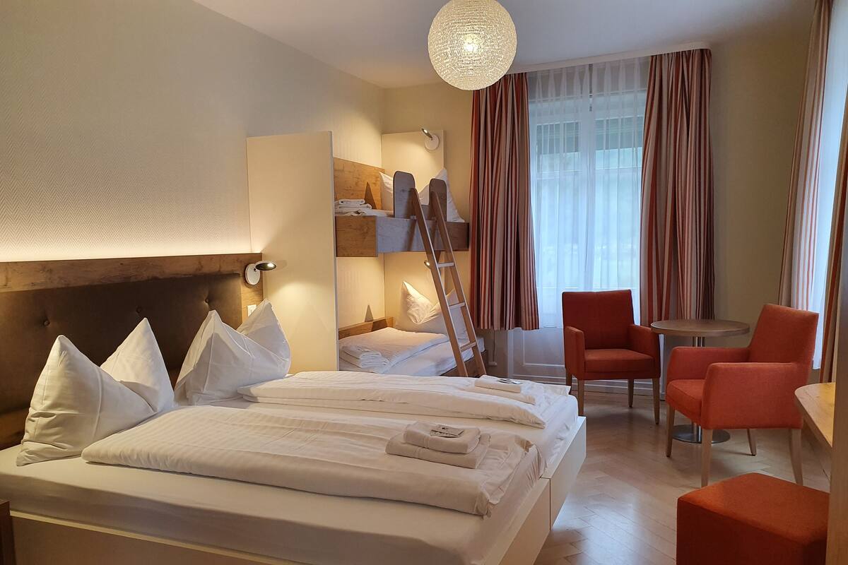 Une chambre d’hôtel avec un grand lit, un bureau, deux chaises, une échelle et une fenêtre avec des rideaux.