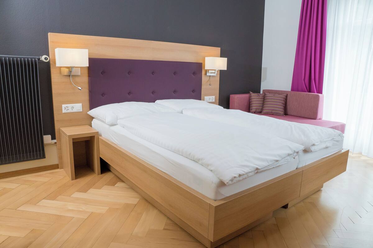Une chambre d’hôtel moderne avec un lit en bois, deux tables de chevet, une tête de lit et un petit coin salon avec un canapé rose.