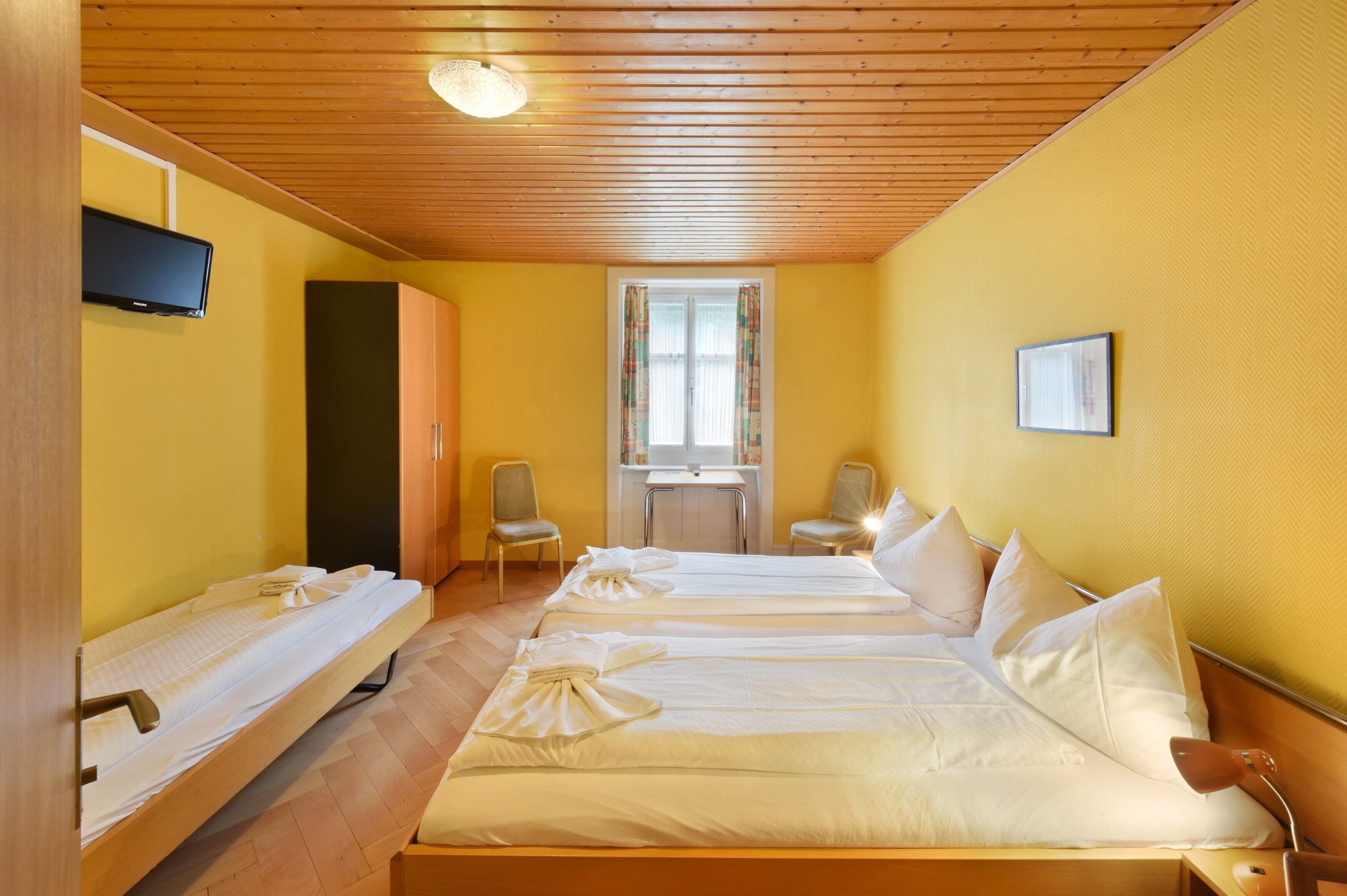 A hotel room with three beds, wooden ceiling, yellow walls, a television, and a window with curtains.