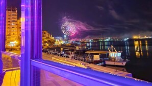 Un paisaje urbano nocturno con fuegos artificiales a lo lejos, un muelle con barcos y edificios con ventanas iluminadas.