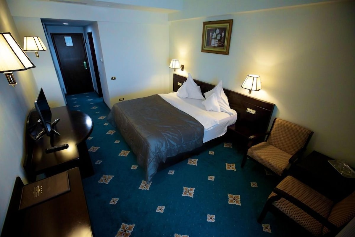 A hotel room with a bed, two bedside lamps, a desk with a lamp, a chair, and a carpet with a geometric pattern.