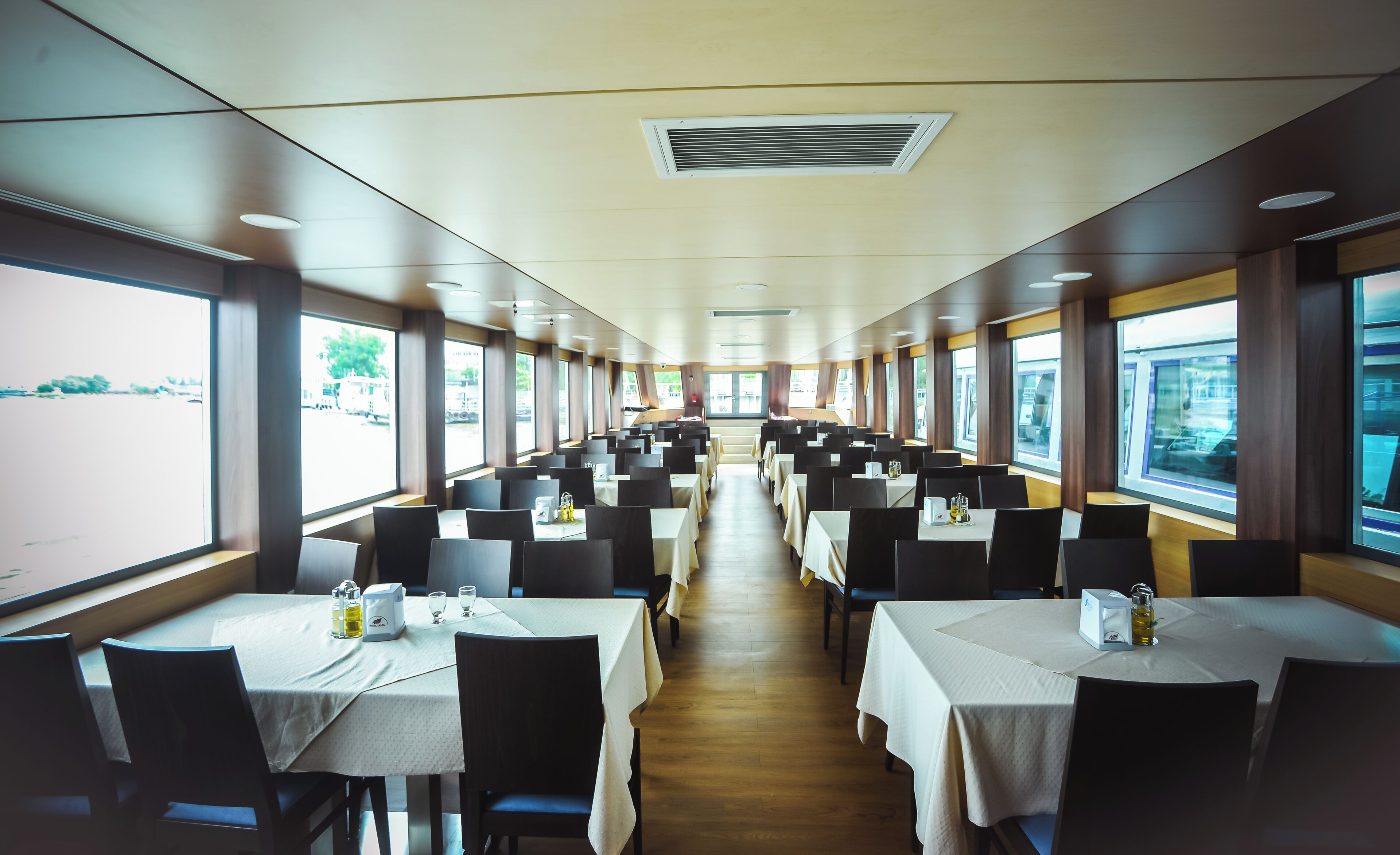 Un restaurant où les tables et les chaises sont disposées en rangées, avec de grandes fenêtres le long des murs et une allée centrale.