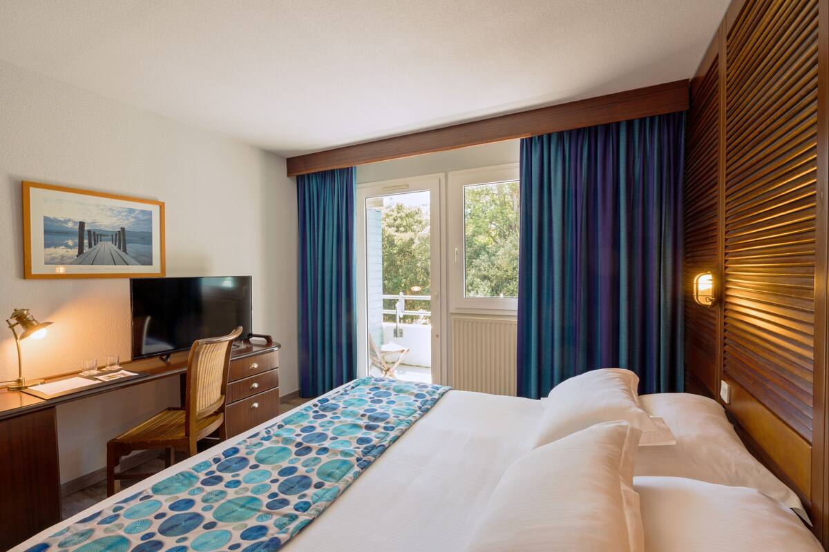 A hotel room with a bed, a desk with a chair, a television, and a window with curtains.