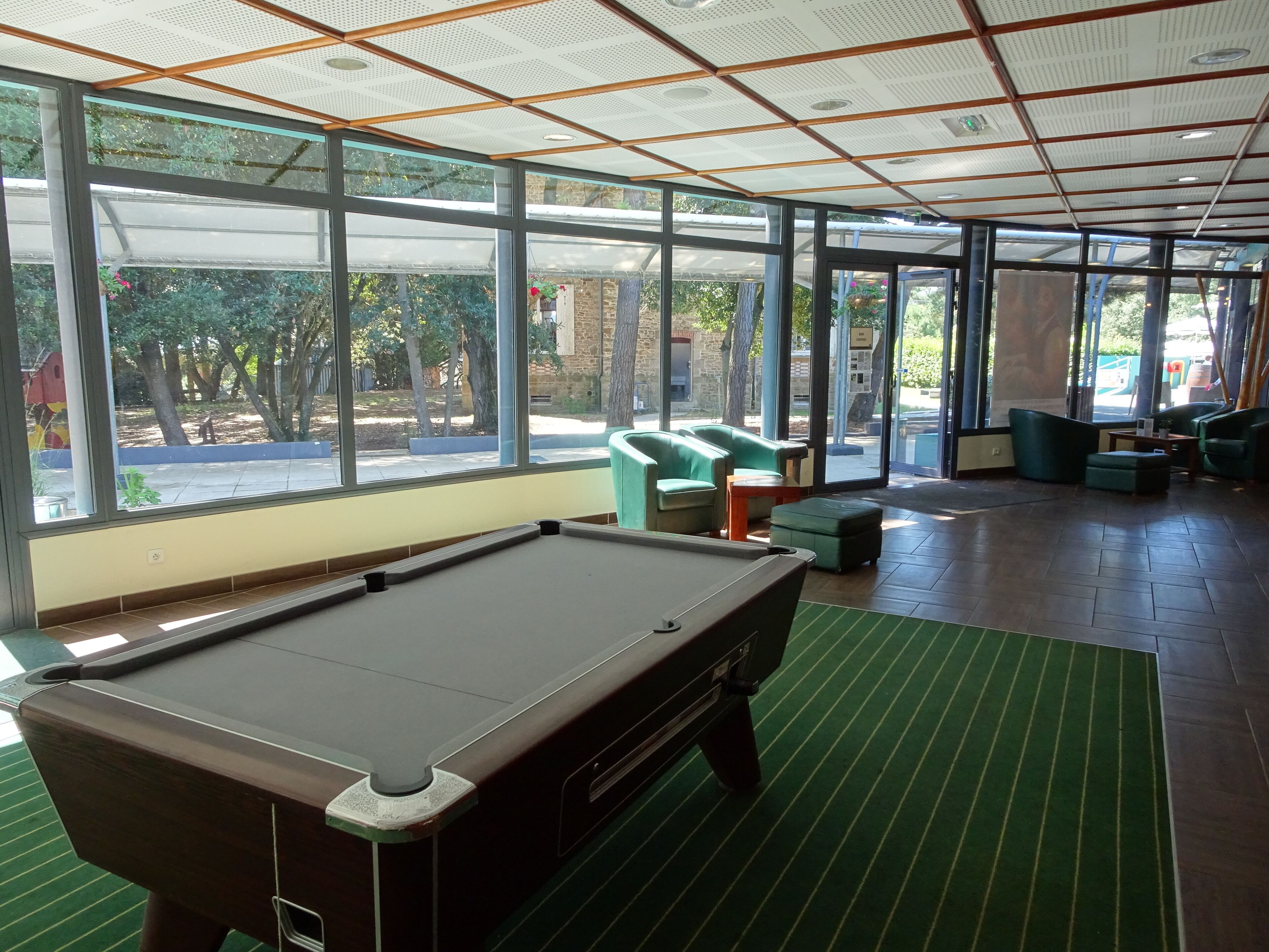 Una mesa de billar en una sala con ventanas amplias, asientos acolchados verdes y vistas al exterior.