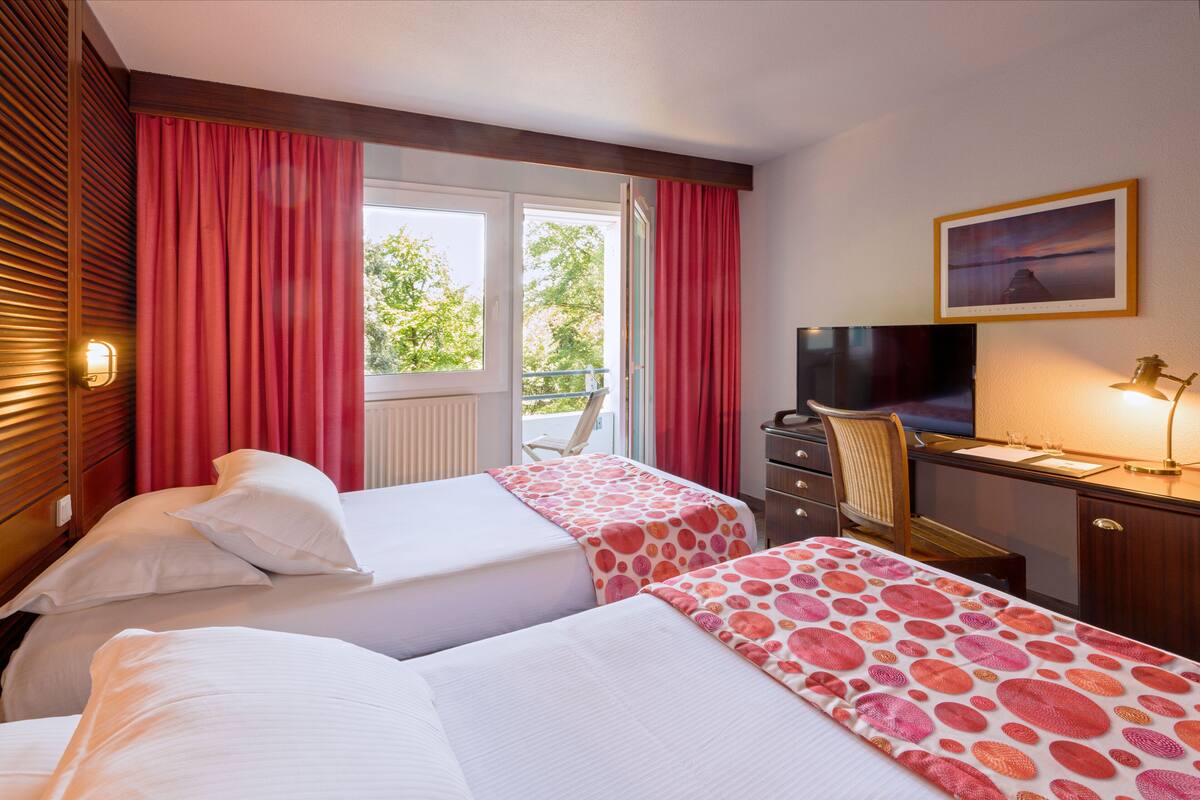 A hotel room with two beds, a desk with a TV, and a window with red curtains.