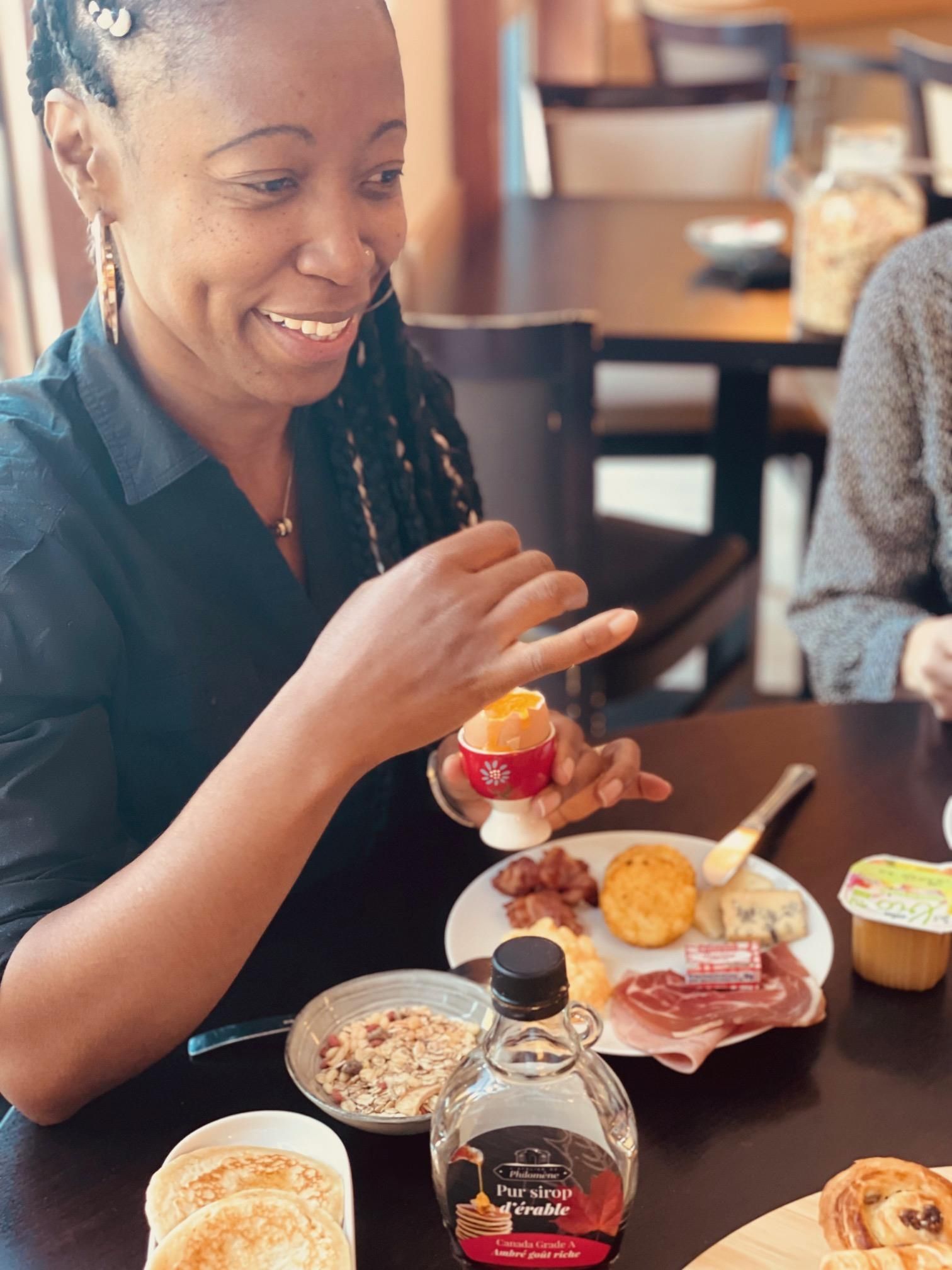 Una persona sentada en una mesa con diversos alimentos para el desayuno, incluyendo tortitas, una botella de jarabe y un plato con carne y queso.