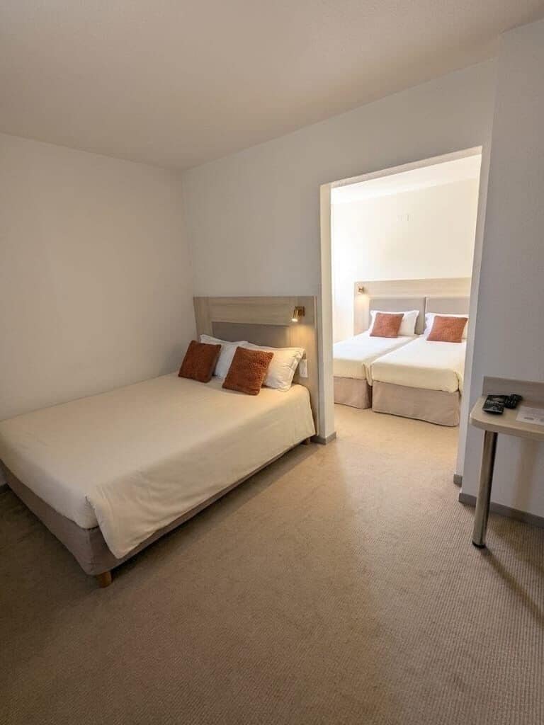 A hotel room with a single bed, a second bed, a small table, and a window.