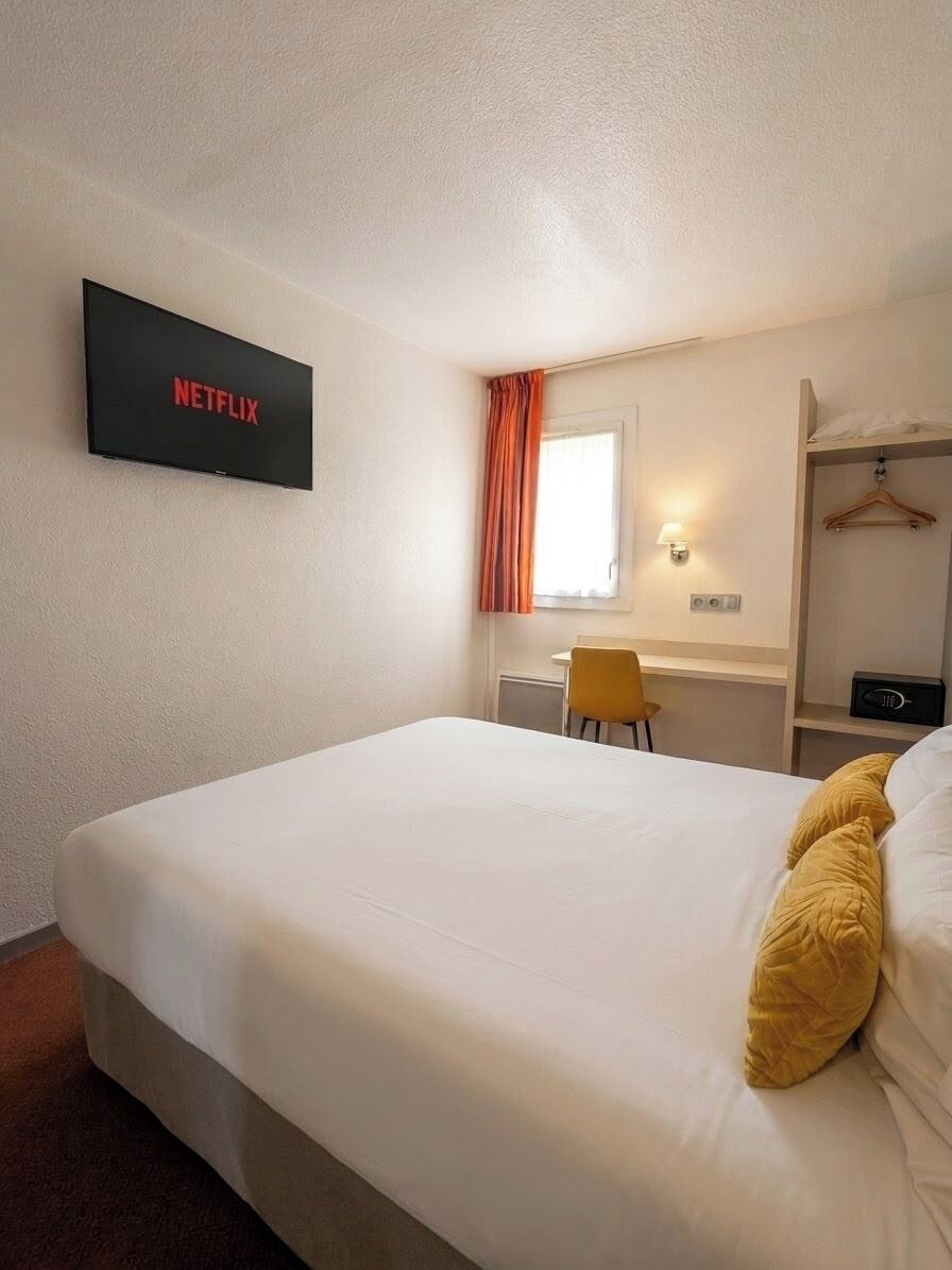 A hotel room with a bed, a desk, a chair, a TV mounted on the wall, and a window with curtains.