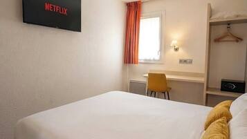 A hotel room with a bed, a desk, a chair, a TV mounted on the wall, and a window with curtains.