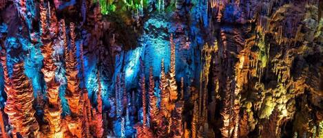 A detailed view of a cave with colorful stalactites and stalagmites, illuminated by artificial lighting.
