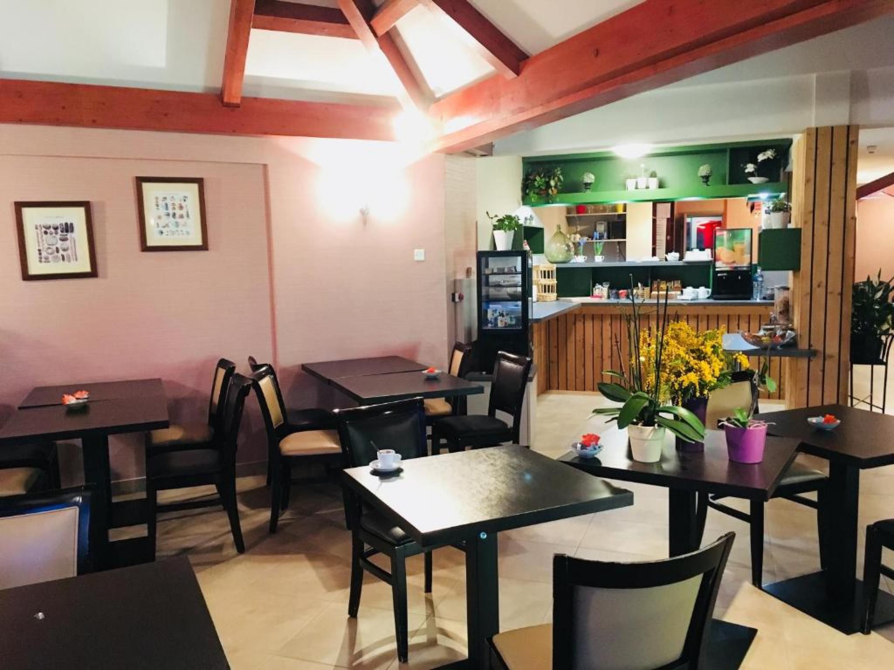 A restaurant with multiple tables and chairs, a counter with a green backdrop, and a visible kitchen area.
