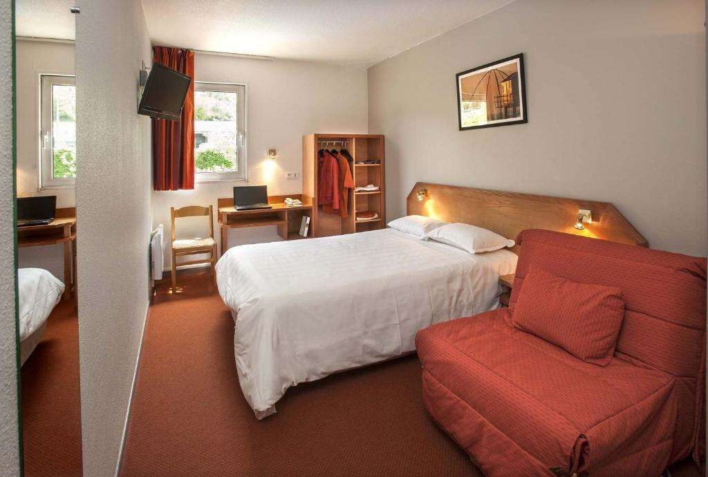 Una habitación de hotel con una cama, un escritorio con una computadora portátil, una silla, un sofá rojo, un armario y un cuadro en la pared.