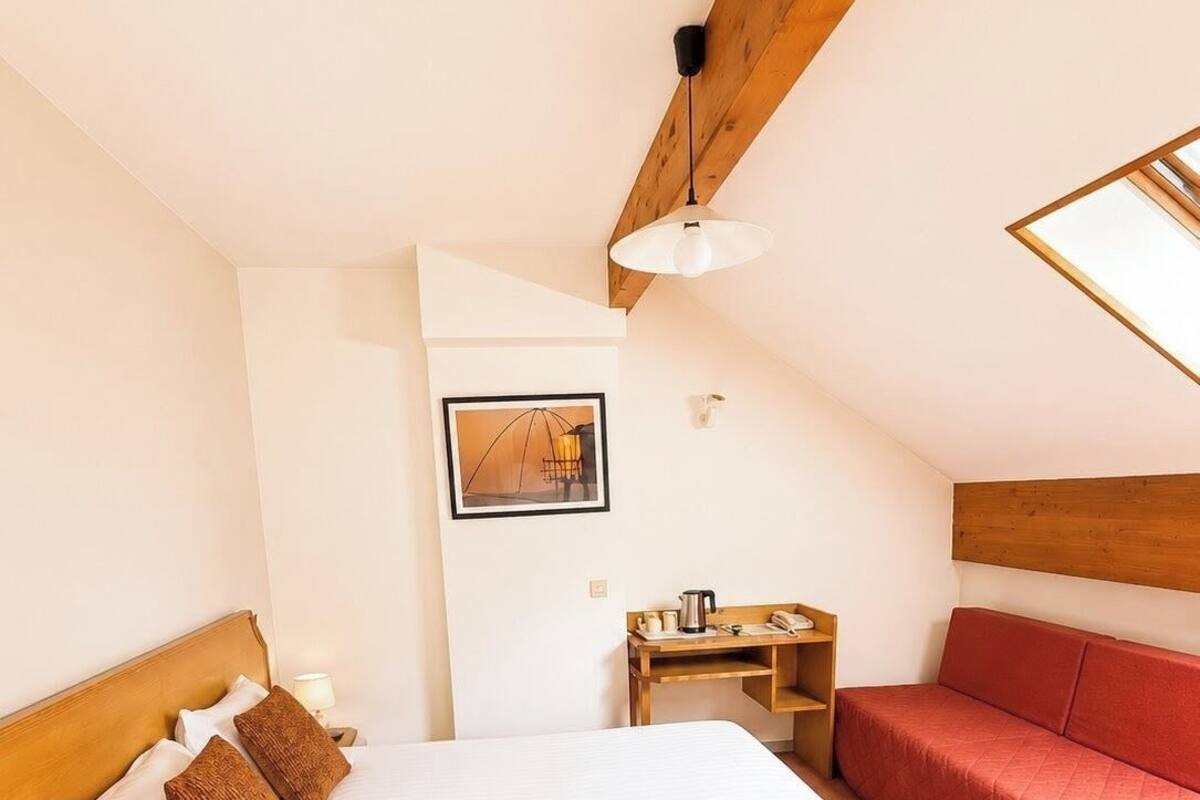 Ein Schlafzimmer mit einem Bett, einem roten Sofa, einem Holznachttisch, einem gerahmten Bild an der Wand und einer schrägen Decke.