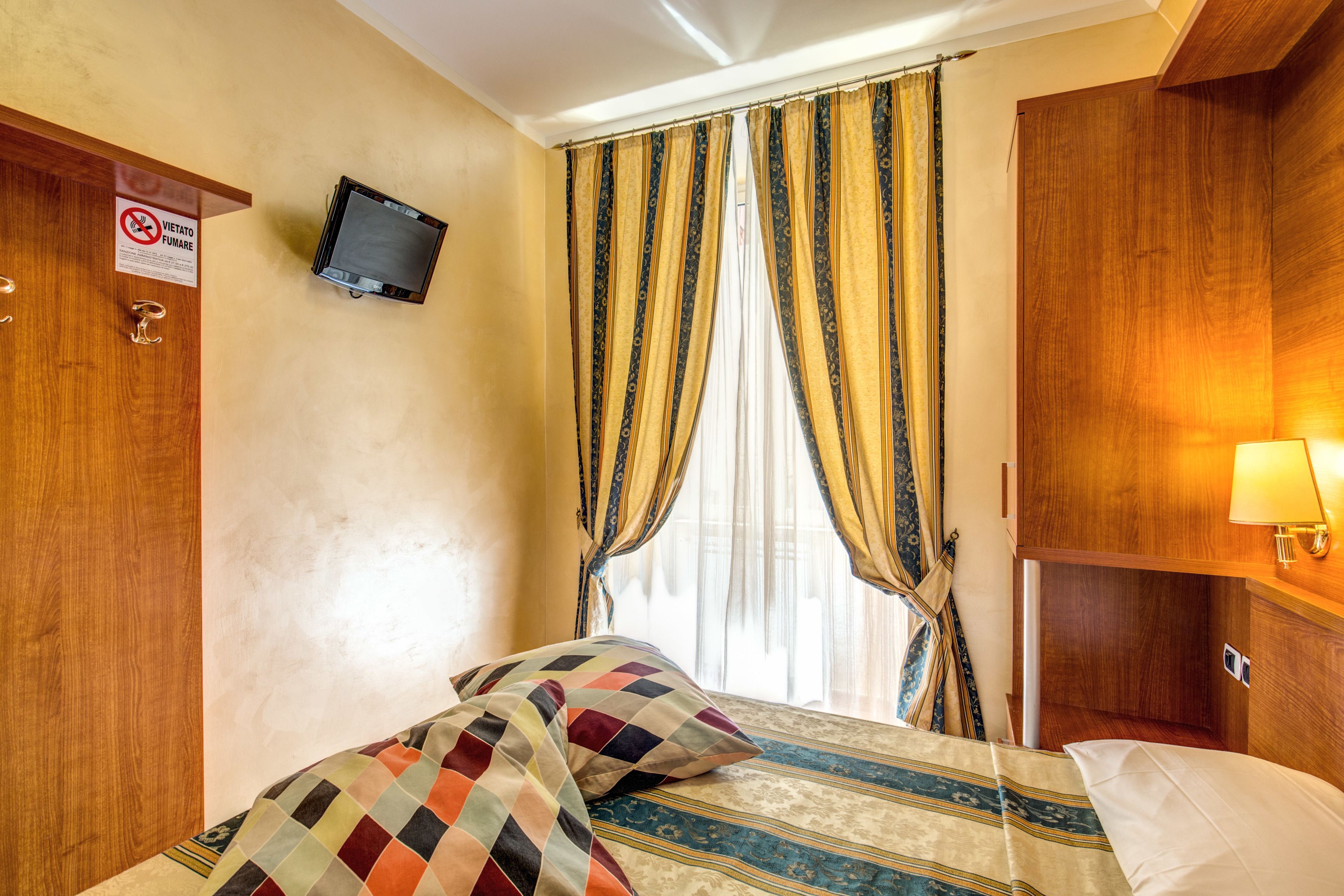 A hotel room with a bed, curtains, a television mounted on the wall, and a wooden wardrobe.