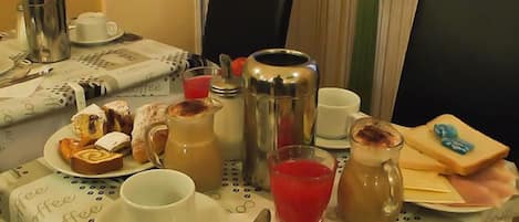 Mesa de café da manhã com cafeteira, bule, xícaras, um copo de suco de uva, doces e um pacote de suco de laranja.