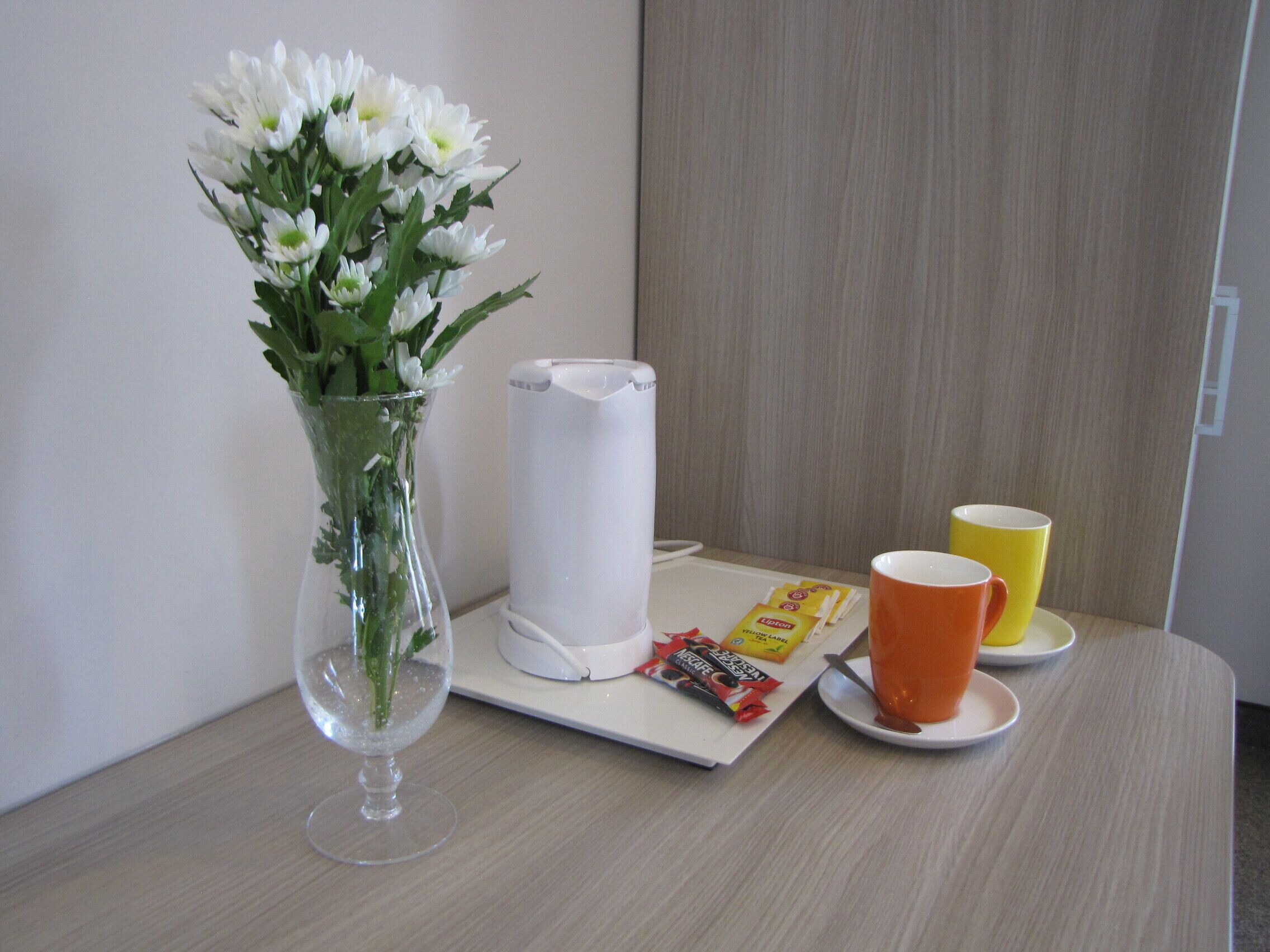 A table with a vase of white flowers, a white electric kettle, a yellow mug, an orange mug, and some snacks.