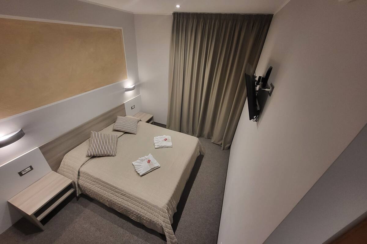 A hotel room with a bed, bedside tables, a TV mounted on the wall, and a window with curtains.