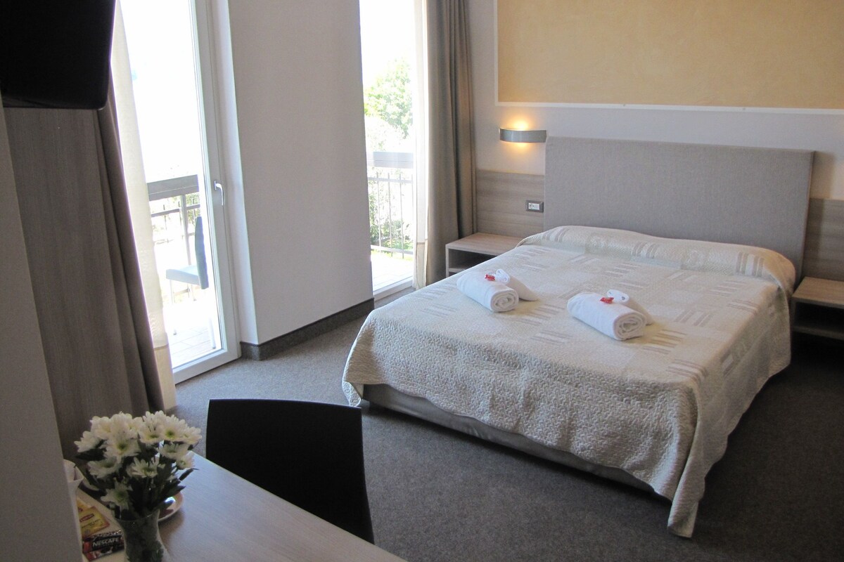 A hotel room with a large bed, a bedside table, a chair, and a view of the outdoors through a sliding door.