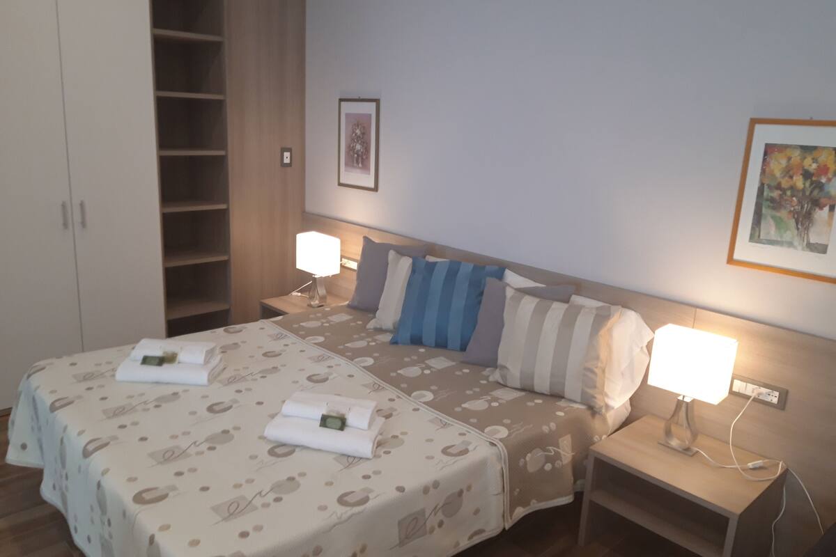 A hotel room with two beds, a wardrobe, and a nightstand with a lamp.