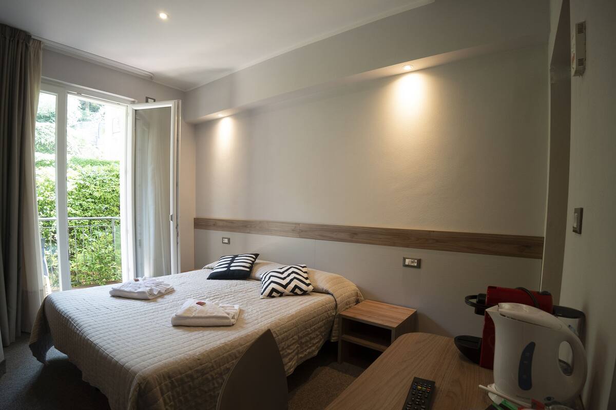 A hotel room with a bed, bedside tables, a desk, a kettle, and a window with curtains.
