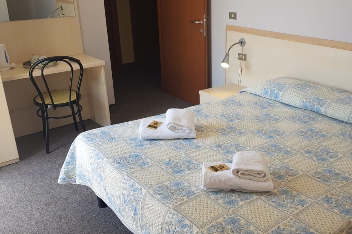 A hotel room with a bed, a desk, a chair, and a nightstand with a lamp.