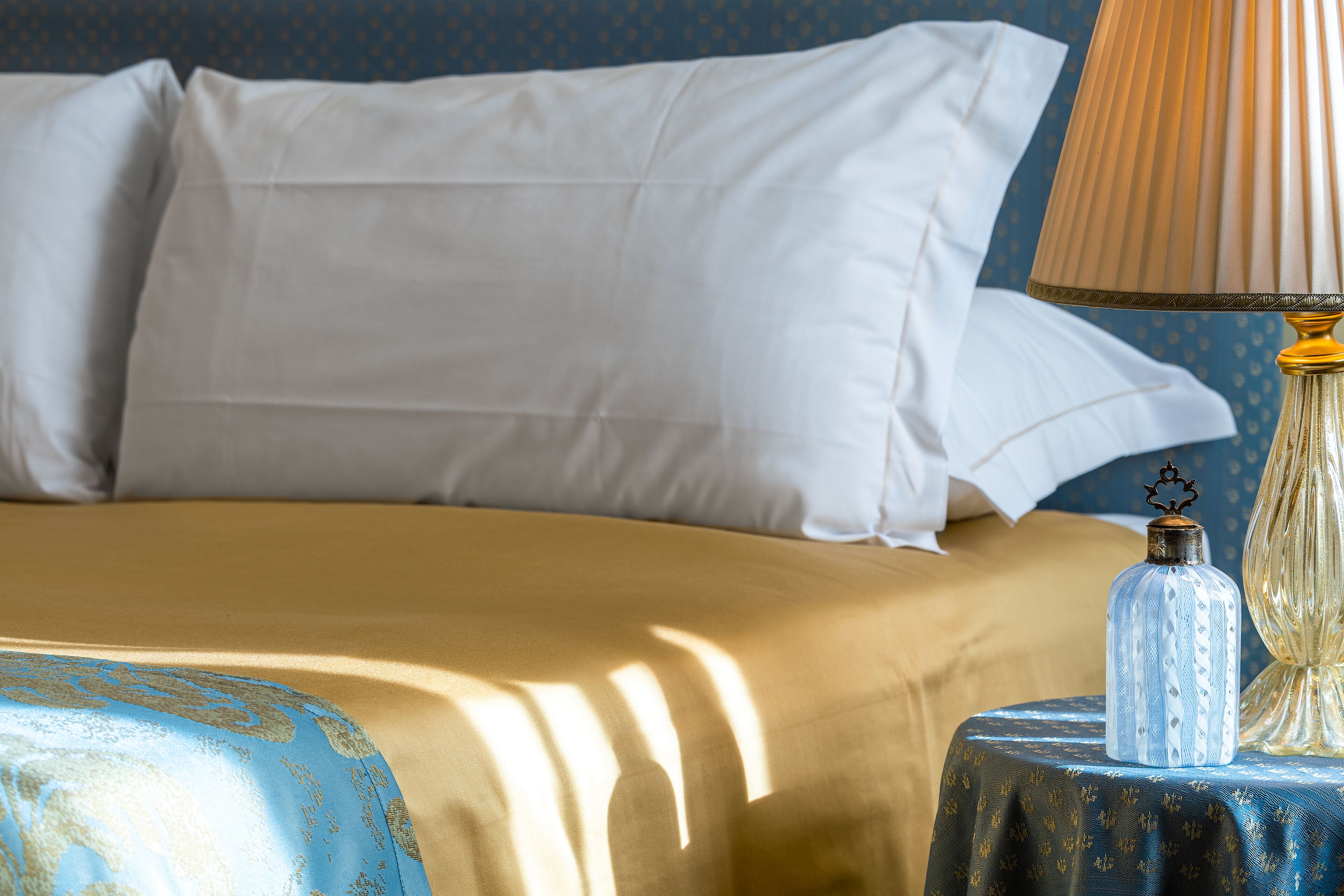 A neatly made bed with white pillows and a gold bedspread, a decorative lamp on a blue table beside it, and a patterned wall in the background.