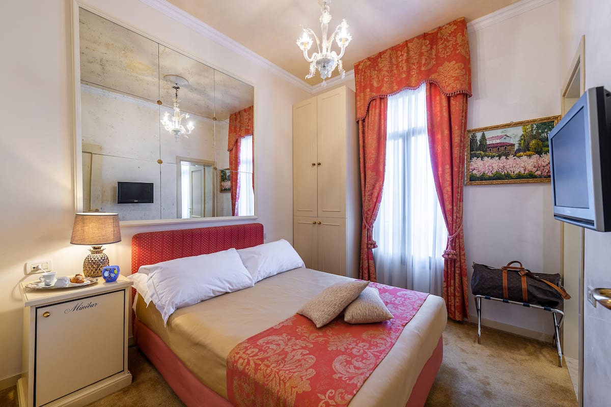 A hotel room with a bed, a bedside table with a lamp, a TV, a wardrobe, and a window with curtains.