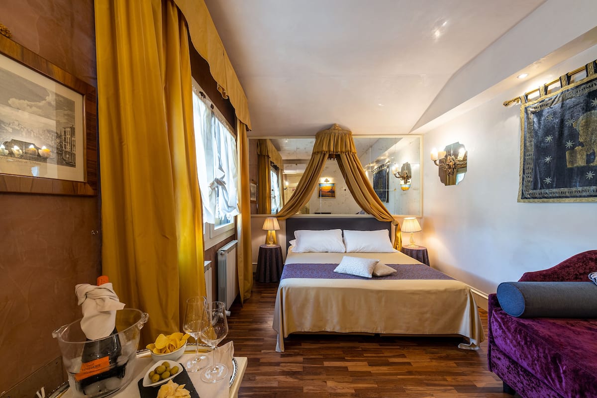 A hotel room with a four-poster bed, a sitting area with a purple sofa, a dining table with a glass of wine, and a bowl of olives.
