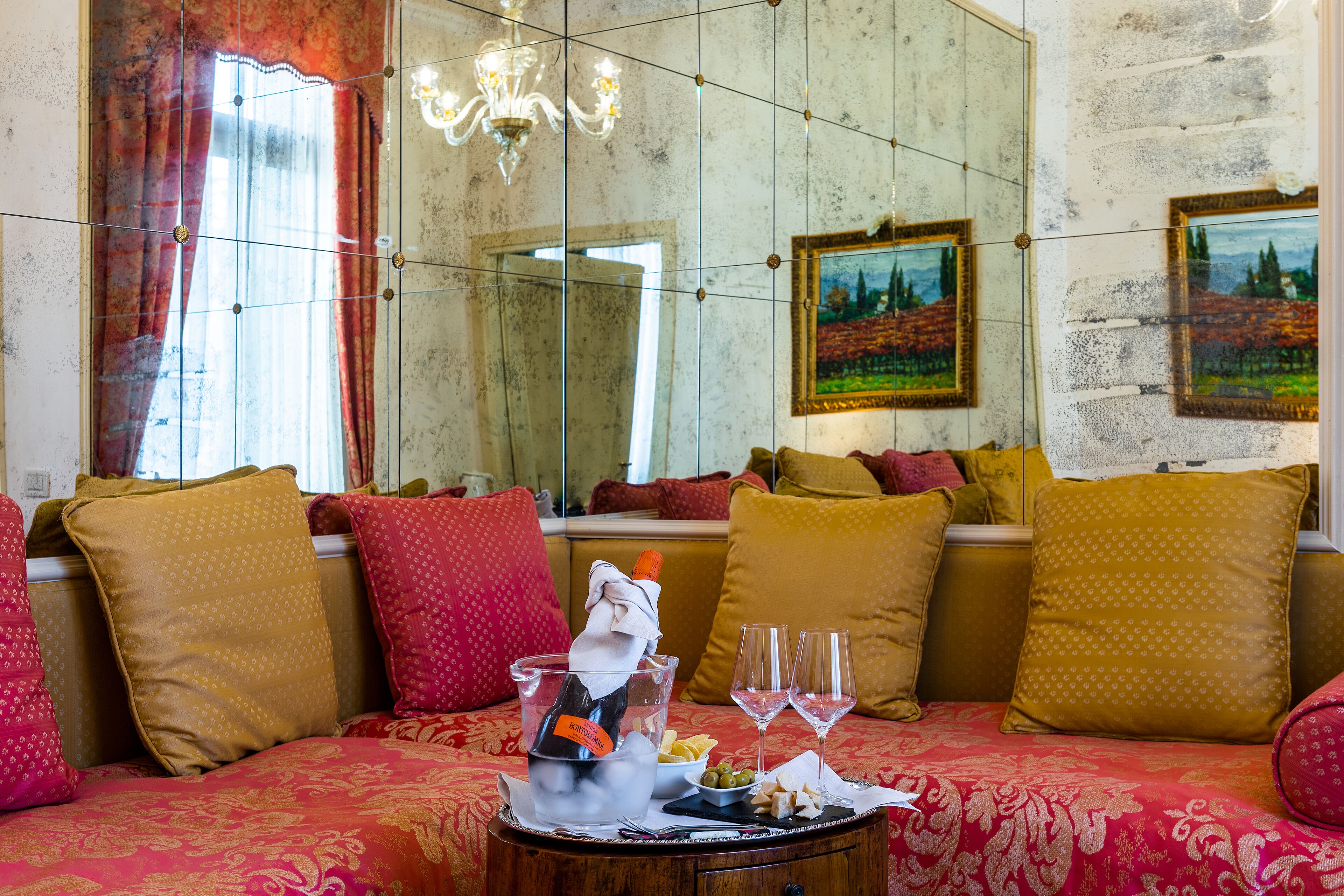 A cozy living room with a sofa, cushions, a table with a bottle and glasses, and a large mirror.