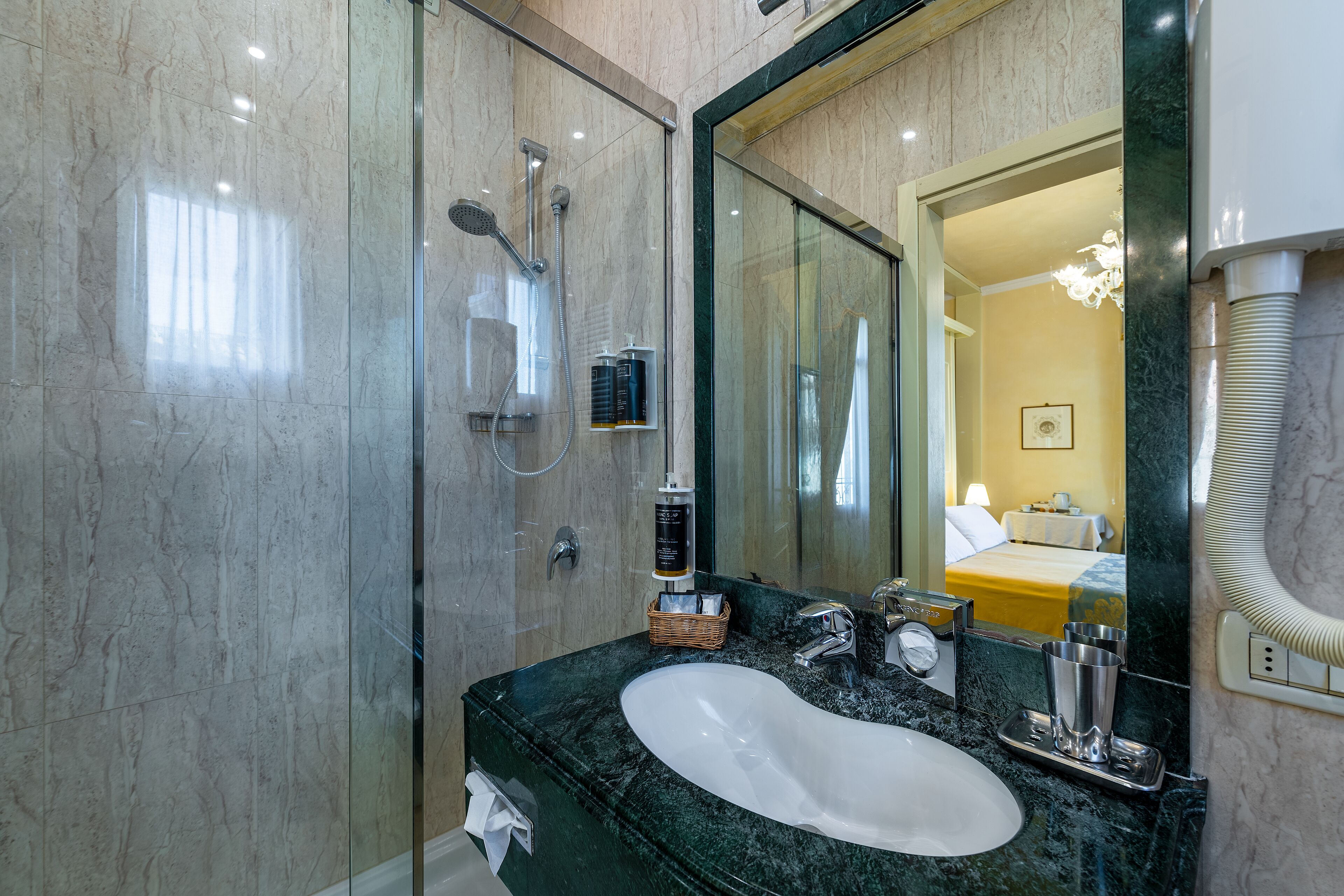 A modern bathroom with a glass shower enclosure, a marble sink, and a large mirror reflecting a bedroom with a bed and a nightstand.